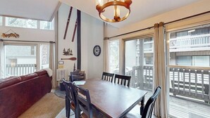 Uma sala de jantar com mesa e cadeiras de madeira, um sofá vermelho, um candelabro e grandes janelas com vista para uma varanda.