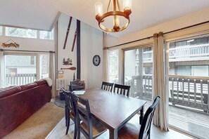 A dining area with a wooden table and chairs, a red sofa, a chandelier, and large windows with a view of a balcony. - Perfect Condo W/ Shared Pools/Hot Tubs, Free WiFi, Full Kitchen & Balcony (Mammoth Lakes)
