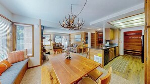 A spacious living area with a wooden dining table, a sofa, and a kitchen in the background. - Inviting condo 500 feet to canyon lodge lifts w/on-site pool, hot tub, sauna (Mammoth Lakes)