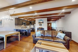 A spacious living area with a leather sofa, wooden coffee table, and a dining table. A kitchen with dark cabinets and a central island is visible in the background. - Sophisticated, dog-friendly condo in nature with fireplace - walk to village (Mammoth Lakes)