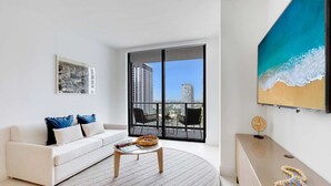 A modern living room with a white sofa, a round coffee table, and a large window with a city view. - Modern 1BR Oasis with Pool & Gym Access (Miami)