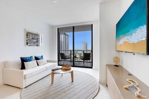 A modern living room with a white sofa, a round coffee table, a large window with a city view, and a painting of a beach scene. - Modern 1BR Oasis with Pool & Gym Access (Miami)