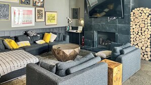 Un salon moderne avec un canapé d’angle gris, une table basse ronde en bois et une cheminée où sont empilés des bûches.