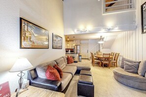Ein Wohnzimmer mit einer Couch, einem Hocker, einem Esstisch und Stühlen. Eine Lampe, ein gerahmtes Bild an der Wand und eine Küche im Hintergrund sind ebenfalls zu sehen.