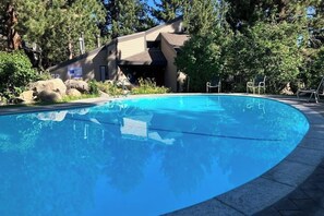 A clear blue pool with a stone border, surrounded by trees and a building with a sloped roof. - Well-equipped mountain-view condo with outdoor pool, hot tub, grill, & fireplace (Mammoth Lakes)