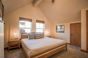 Un dormitorio con una cama grande, dos ventanas con persianas, una mesita de noche con lámpara, una puerta y un cuadro enmarcado en la pared.