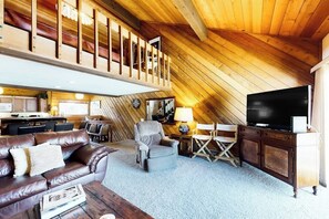 Ein Loft-Wohnzimmer mit hohen Decken, Holzbalken, einem Ledersofa, einem Fernseher und einem Essbereich mit einer Küche im Hintergrund.