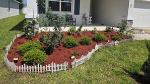 Ein Wohnhaus mit Veranda, weißem Zaun und einem angelegten Garten mit Mulch und verschiedenen Sträuchern.