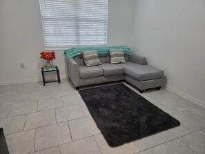 Un salon avec un canapé d’angle gris, un tapis noir, une petite table sur laquelle est posé un vase de fleurs et une fenêtre avec des stores.