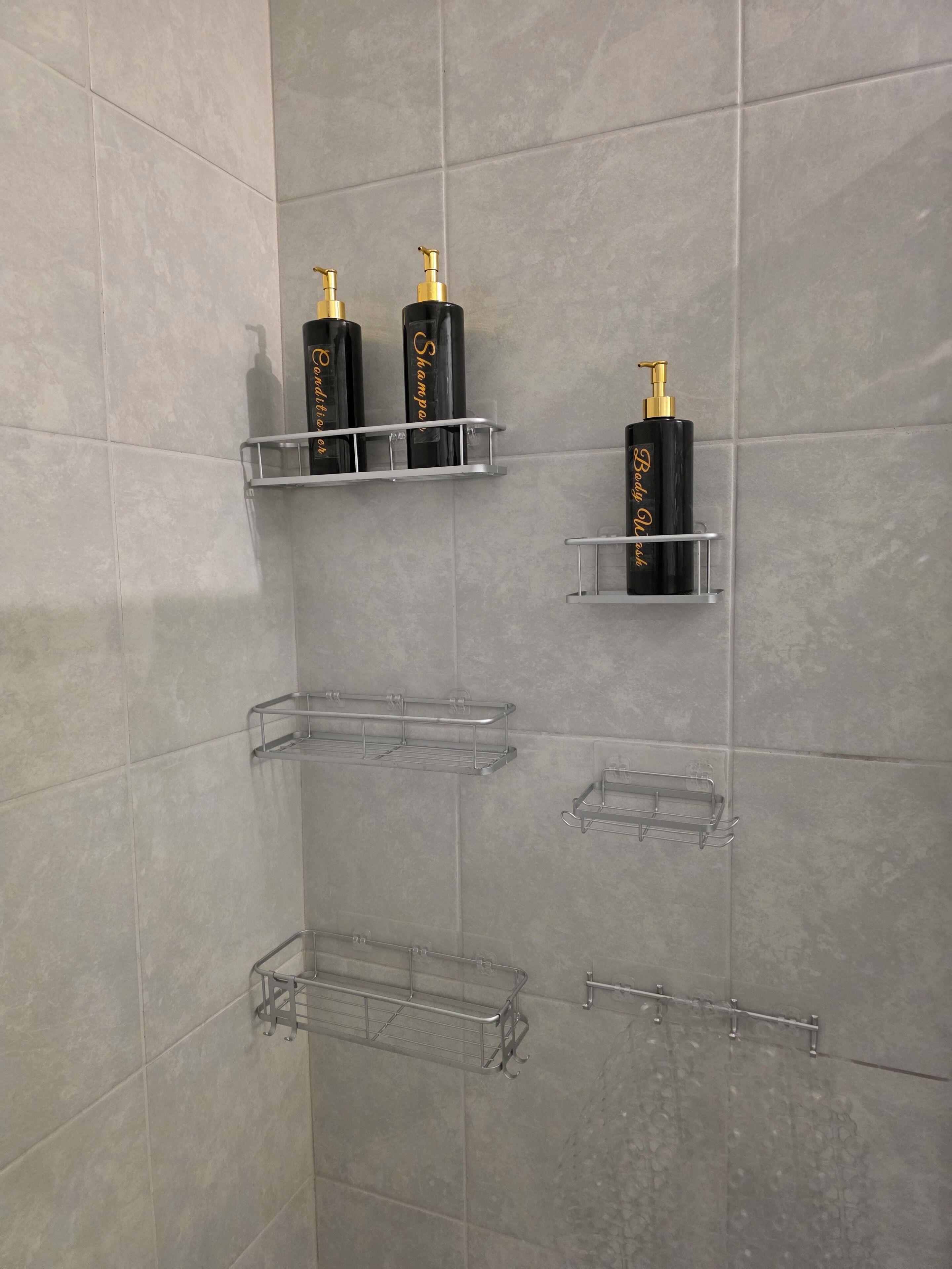 A bathroom with grey tiles, multiple clear plastic shelves, and black bottles with gold pumps.