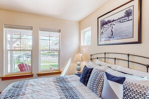 A bedroom with a bed, two windows, a lamp, and a framed picture on the wall.