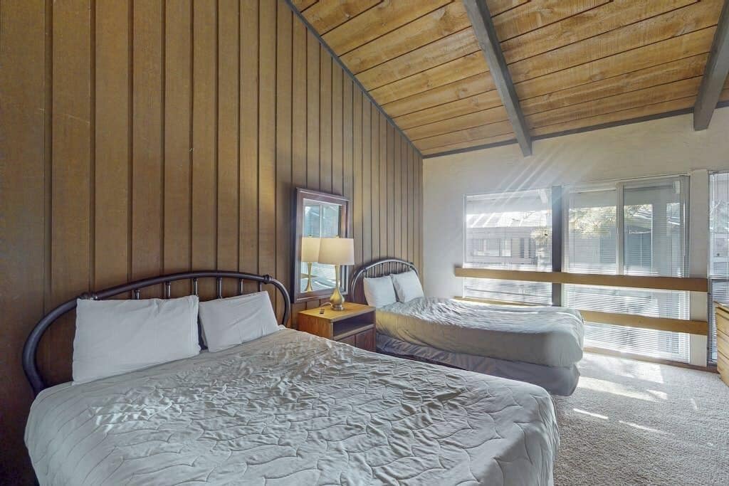 A bedroom with two beds, wooden panel walls, a window with blinds, and a nightstand with a lamp.