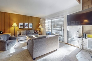 Ein modernes Wohnzimmer mit einer Couch, Sesseln und einem Couchtisch. An der Wand befinden sich Bilder in Rahmen, eine Lampe und eine Glastür zu einem Balkon.