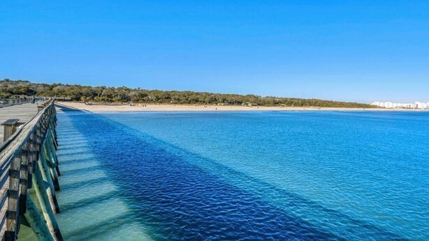 Un molo che si protende in acque cristalline blu, con una spiaggia sabbiosa e vegetazione sullo sfondo.