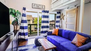 Zona giorno con divano blu, tavolino, tende a righe e un cartello con la scritta 'GATHER'.