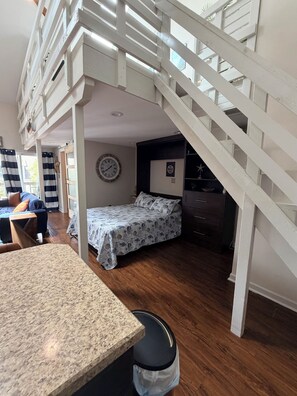 A bedroom with a bed, a clock, a 'No Wake Time' sign, a staircase, and a dining area with a table. - Surfside Studio + Loft -Arcade Room, Pool, Balcony, Golf Cart Friendly (surfside beach)