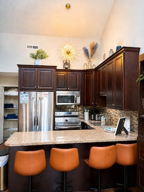 A kitchen with dark wood cabinets, stainless steel appliances, a granite countertop, and three orange bar stools. - Surfside Retreat, Arcade, Pool, Hot Tub & Tennis (surfside beach)