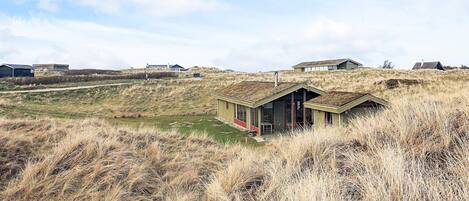 A remote, single-story building with a sloped roof, surrounded by tall grass and dunes.