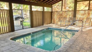 A covered outdoor swimming pool with a tiled deck, a metal ladder, and a view of a vehicle through the fence.
