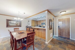 A dining area with a wooden table and chairs, a kitchen with wooden cabinets, and a stone-tiled floor. - Cozy condo w/ shared pool, hot tub & sauna, near outdoor activities (Mammoth Lakes)