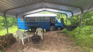 A covered outdoor area with a blue bus, white plastic chairs, and a fire pit. - Peaceful 1-bedroom caravan with AC in Mammoth Spring. (Mammoth Spring)