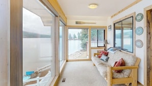 Una habitación con un amplio ventanal corredera que da a un lago, un banco de madera con cojines y una pared decorada con cuadros.