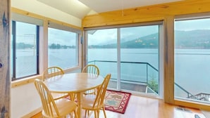 Una habitación con una mesa y sillas de madera, ventanas amplias con vistas al agua y colinas, y una barandilla de cristal.