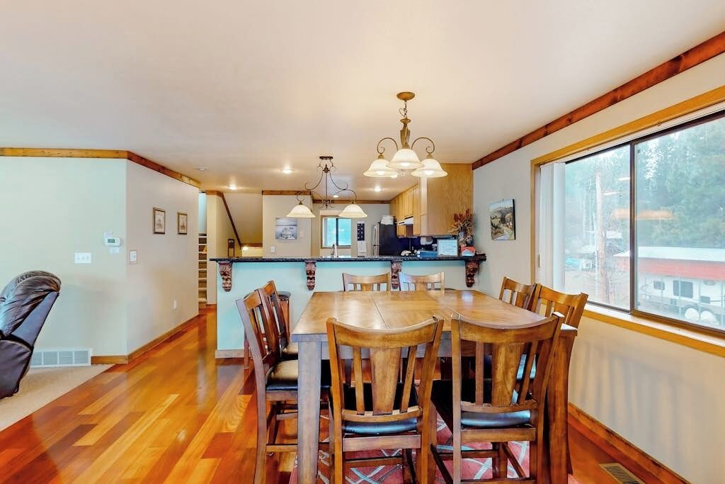A dining area with a wooden table and chairs, a kitchen in the background, and a large window with a view.