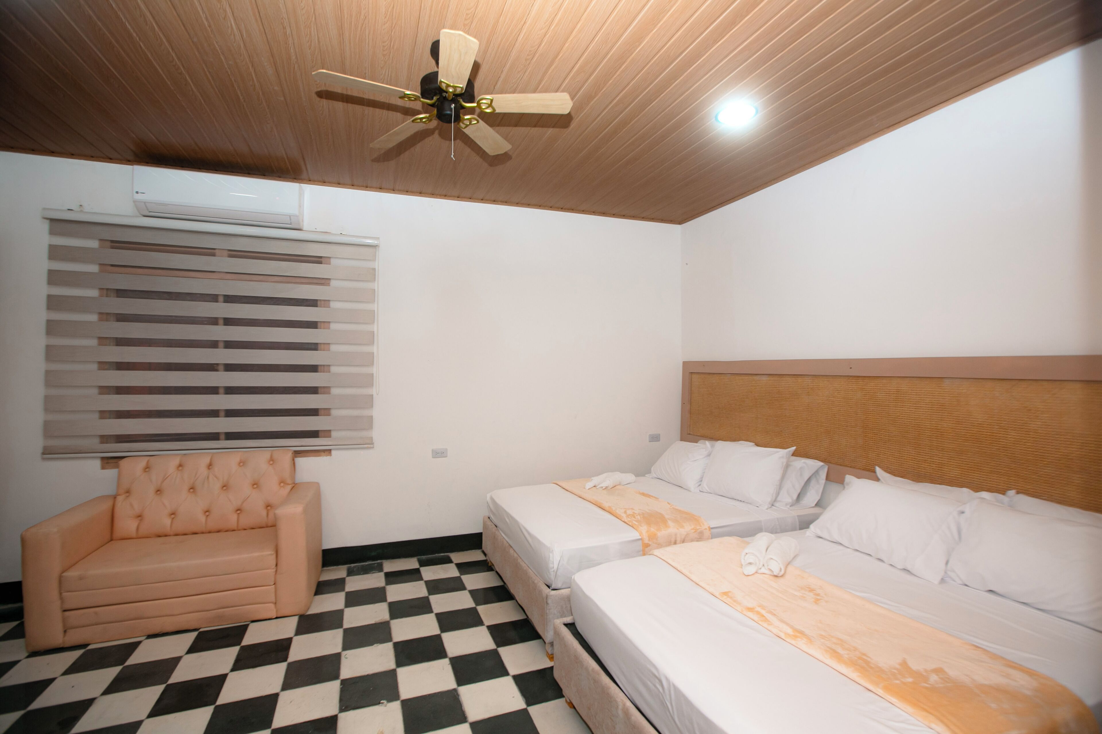 Habitación de hotel con piso a cuadros, dos camas individuales, un sofá, un ventilador de techo y una ventana con persianas.