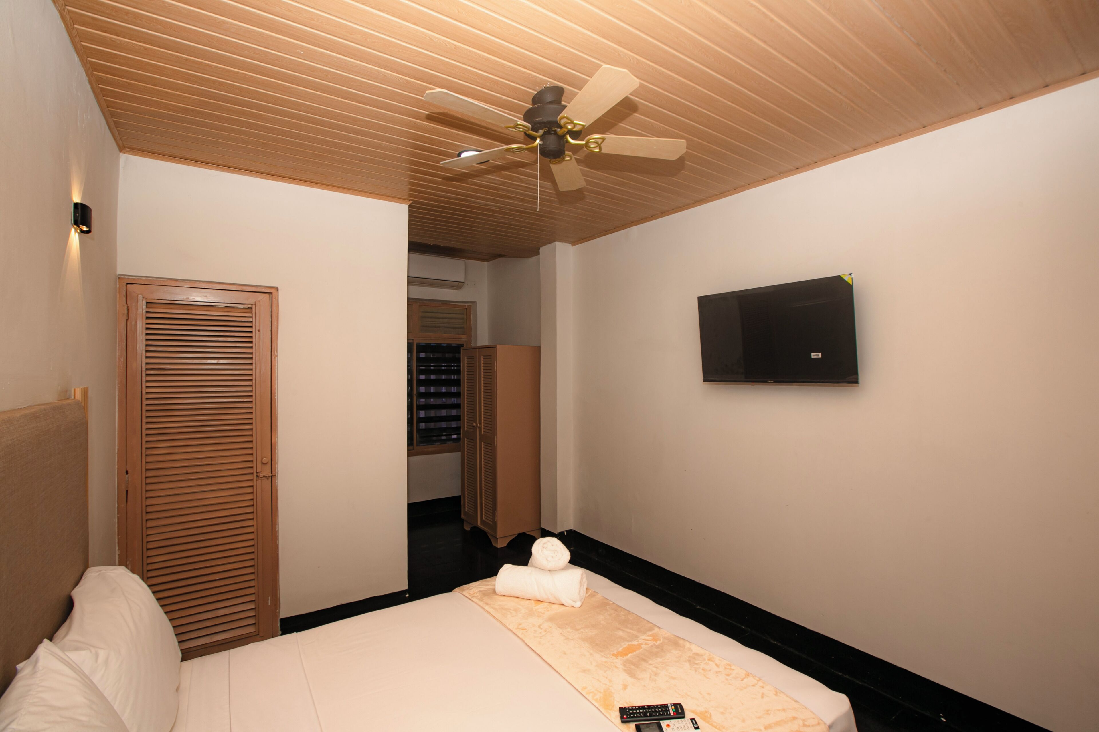 A hotel room with a bed, a ceiling fan, a television, and a wooden wardrobe.