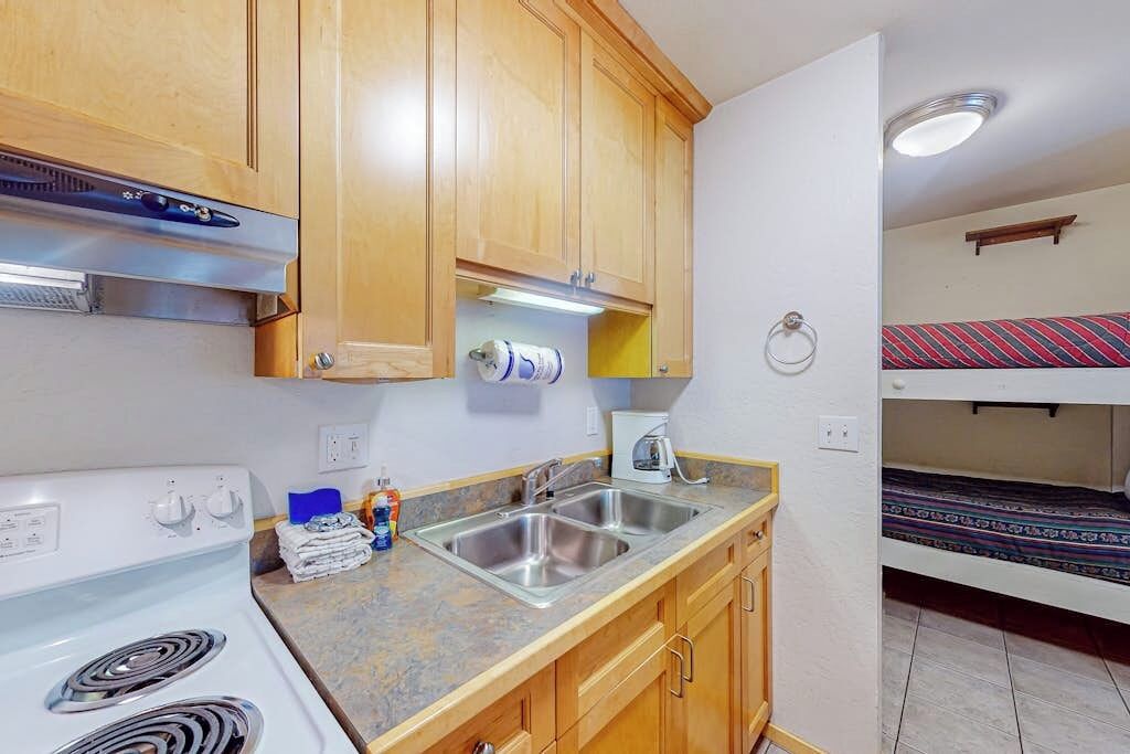 Une cuisine compacte avec des meubles en bois, une cuisinière blanche et un évier. Dans la pièce adjacente, on trouve un lit superposé avec une couverture à rayures.