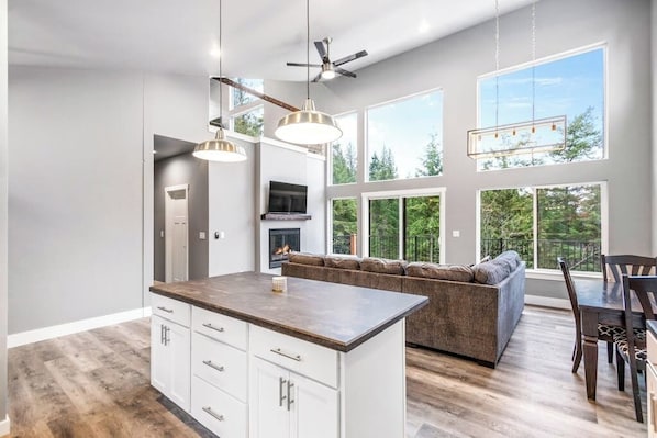 A modern kitchen with a central island, a dining area with a table and chairs, a built-in fireplace, and large windows offering a view of the outdoors.