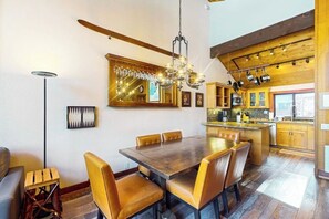 Ein Essbereich mit Holztisch und -stühlen, einer Kristallleuchtre und einer Küche mit Holzschränken und hängenden Töpfen.