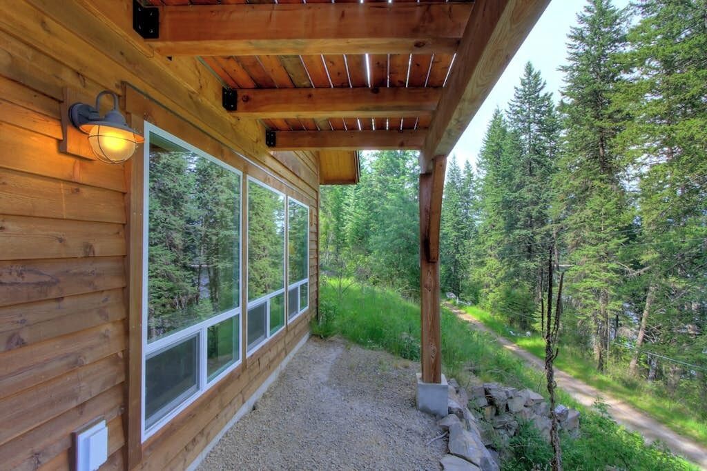 Une cabane en bois dotée de grandes fenêtres, d’un porche abrité et entourée d’un paysage boisé.