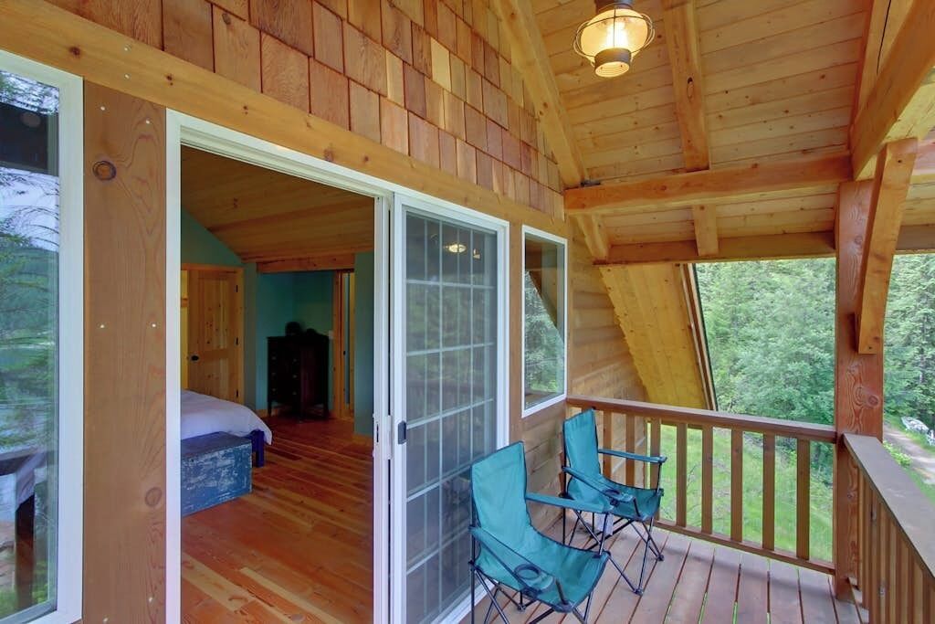 A wooden cabin with a balcony, a bedroom with a bed and a blue chair, and a view of the outdoors.