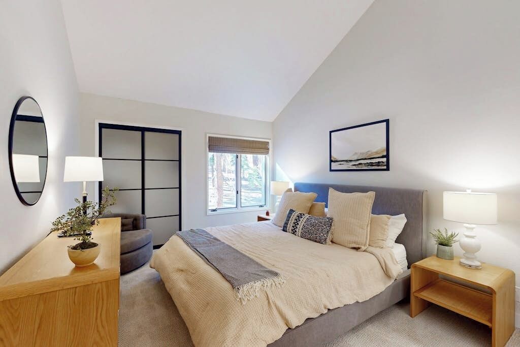 A bedroom with a bed, bedside tables, a mirror, a window, and a wall-mounted artwork.