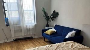 A bedroom with a bed, a blue sofa, a potted plant, and a window with sheer curtains.