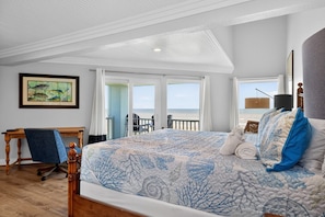 Ein Schlafzimmer mit einem großen Bett, einem Schreibtisch, einem Stuhl und Blick auf den Strand, sowie einem Bild an der Wand.