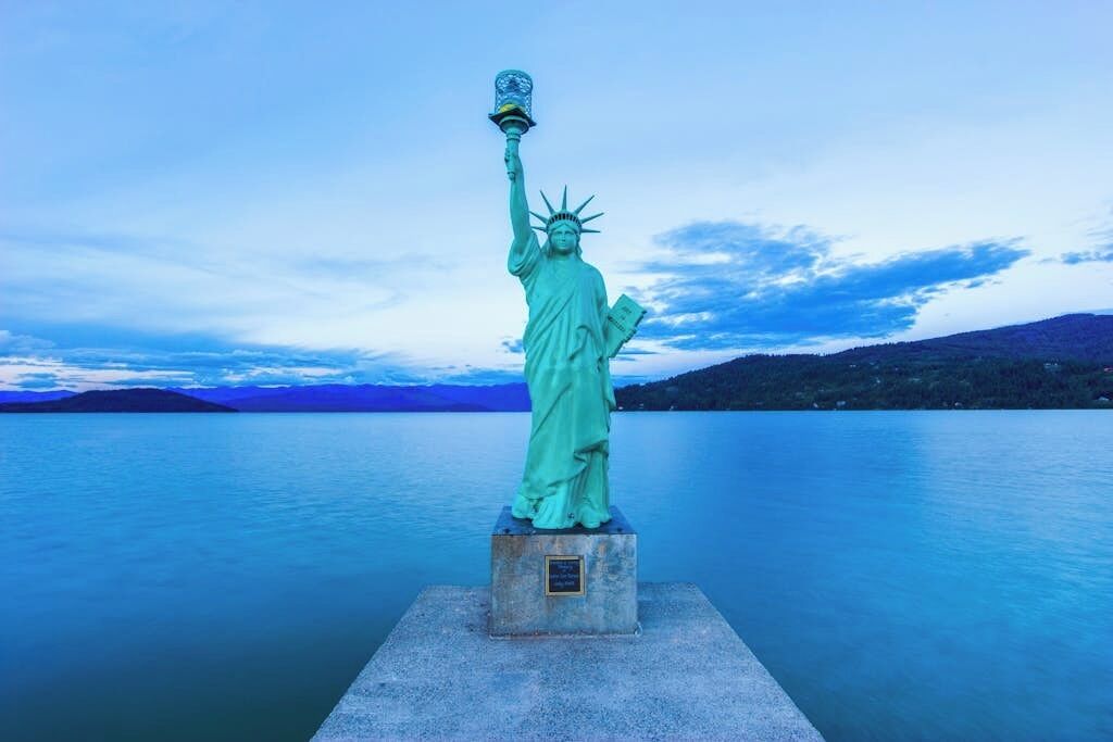 Eine Statue einer Frau, die eine Fackel hält, steht auf einer Plattform vor einem See und Bergen.