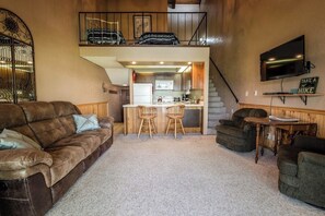 A cozy living room with a brown leather couch, two wicker chairs, a small wooden table, a TV mounted on the wall, and a staircase leading to an upper level. - Waterfront condo w/ complimentary boat slip, shared pool, & sports courts (Sandpoint)