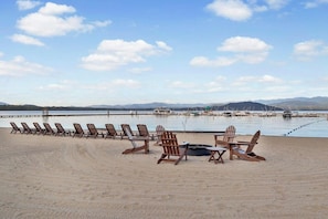 A beach with wooden chairs and tables arranged for outdoor dining, a body of water, and distant mountains.