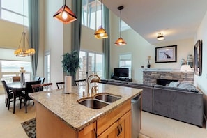 A modern kitchen with granite countertops, a double sink, pendant lighting, a dining area with a table and chairs, a gray sofa, a fireplace, and a large window.