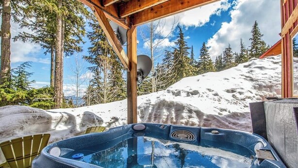 Een hottub in een besneeuwde omgeving, met een houten constructie erboven, omringd door bomen en een met sneeuw bedekte berg.