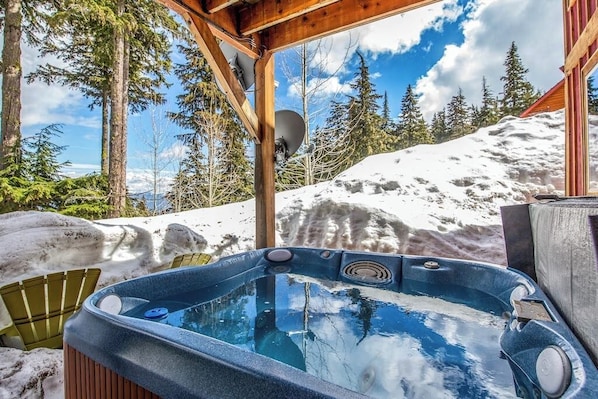 Ein Whirlpool in verschneiter Winterlandschaft, überdacht von einer Holzhütte, umgeben von Bäumen und einem schneebedeckten Berg.