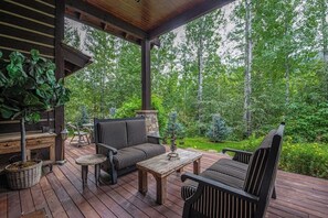Eine Holzterrasse mit Gartenmöbeln, einem Kamin aus Stein und Blick auf einen bewaldeten Bereich.