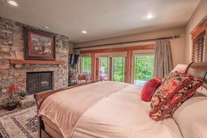 Ein Schlafzimmer mit Kamin aus Stein, einem Bett mit beiger Bettdecke, einem roten Kopfkissen und Blick ins Grüne durch große Fenster.
