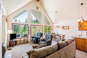 A spacious living area with a high ceiling, large windows, a fireplace, and a kitchen area.