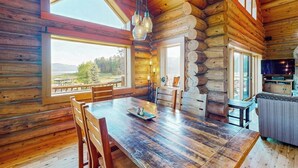 En stuga byggd av timmer med en matplats, stolar, ett köksområde och stora fönster som ger utsikt över omgivningen.