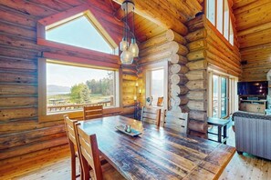 Ein Holzblockhaus-Interieur mit Esstisch, Stühlen, einer Küchenzeile und großen Fenstern mit Blick ins Freie.