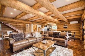A log cabin interior with a large leather sectional sofa, a wooden coffee table, and exposed wooden beams.